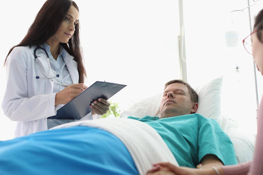 A female doctor stands over a male patient with eyes closed, and his wife holds his hand. Medical malpractice is one cause of wrongful death lawsuits that can provide families with wrongful death compensation.