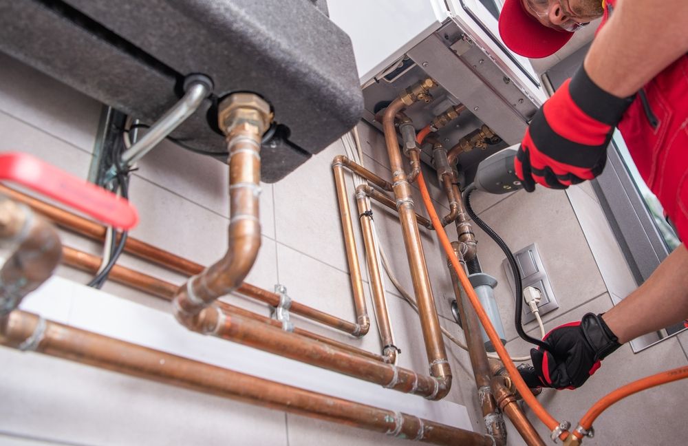 Technician in work gloves servicing the copper gas and water lines beneath a wall-mounted heater