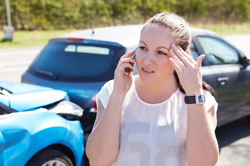 A woman making a phone call, standing in front of two vehicles damaged in a rear-end collision; notifying authorities is one of the first steps to take after an Uber accident.
