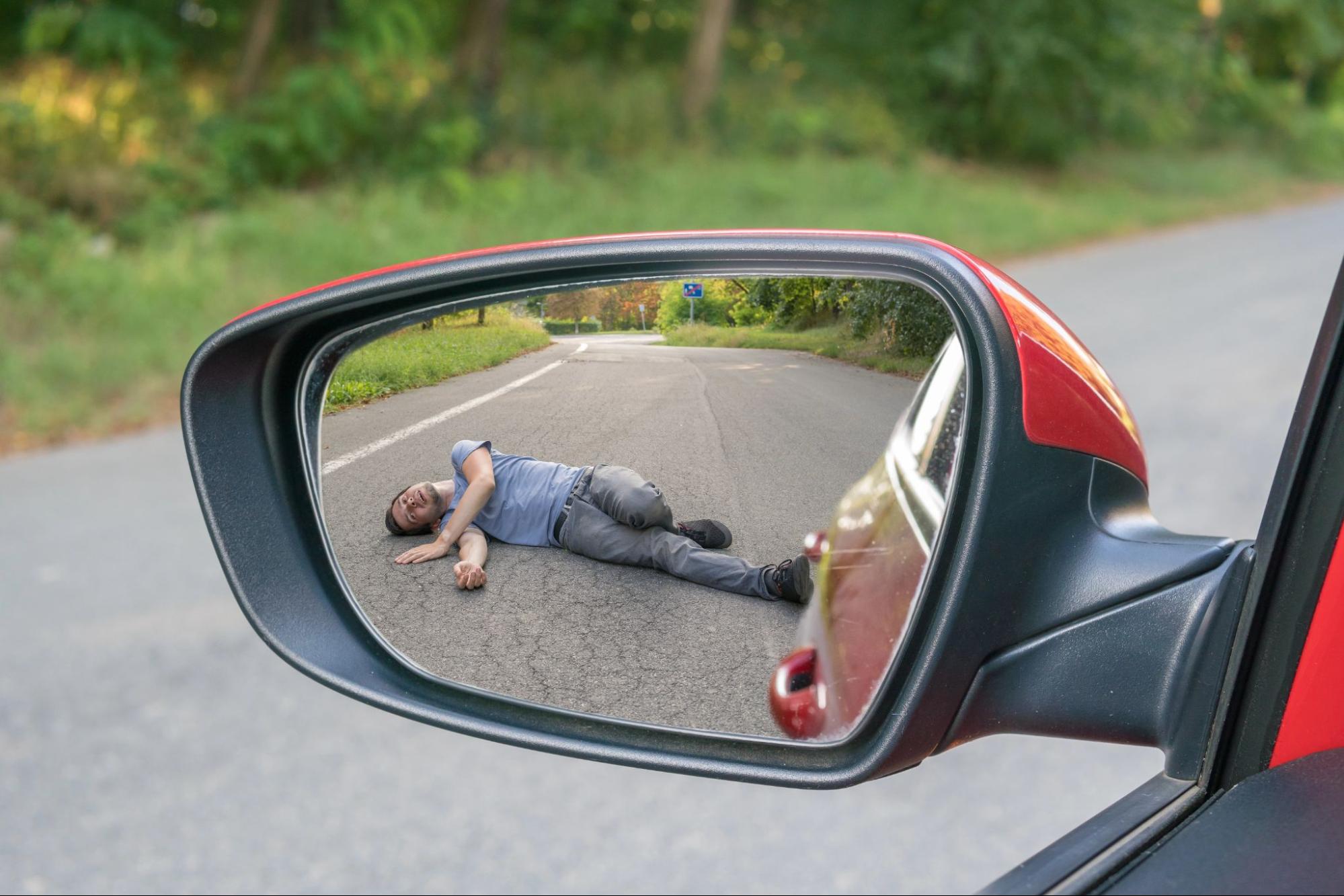 A sideview mirror captures an injured man lying on the road as a red car drives away.