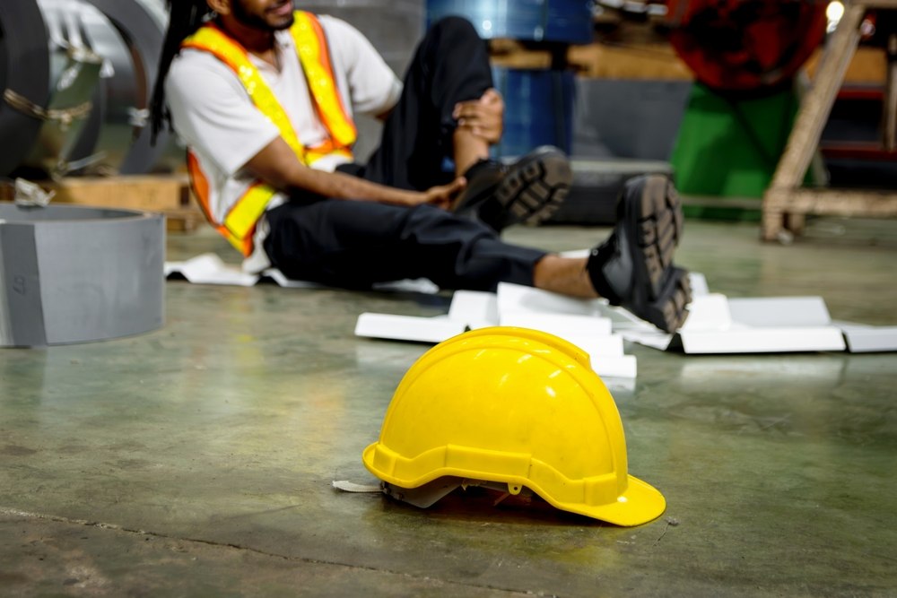 Warehouse worker sitting on the floor holding his ankle after an injury, with a hard hat lying nearby