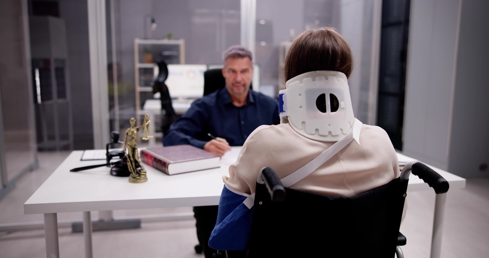 Injured worker in a wheelchair wearing a neck brace and arm sling consulting with an attorney at a desk