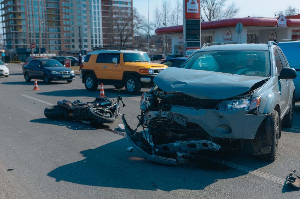 SUV with severe front-end damage and a downed motorcycle after a car accident on a narrow city street