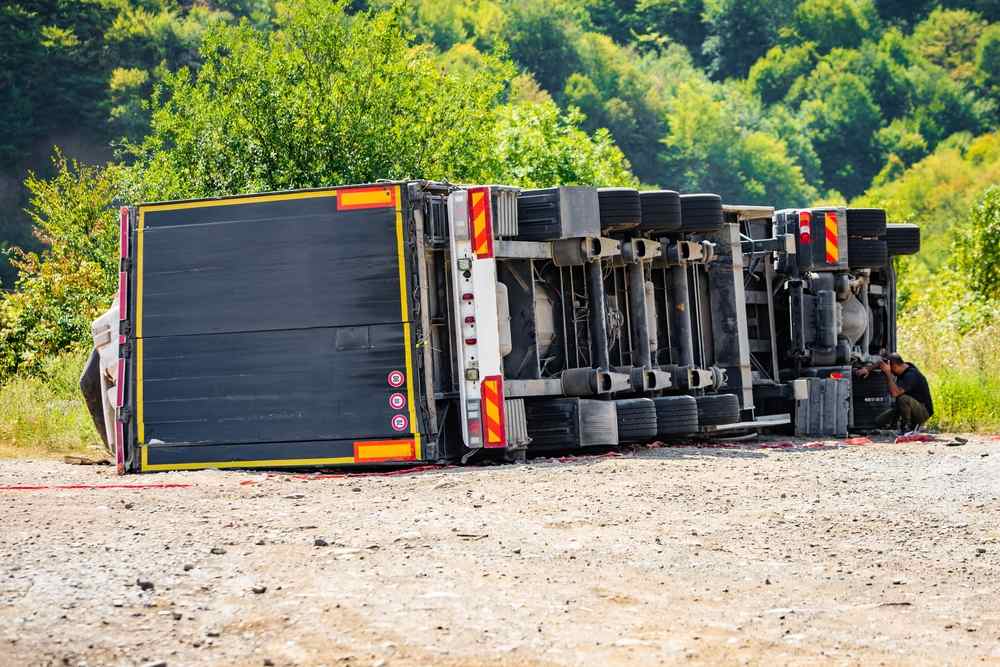 Overturned semi trailer lying on its side along a gravel shoulder while an investigator photographs the underside