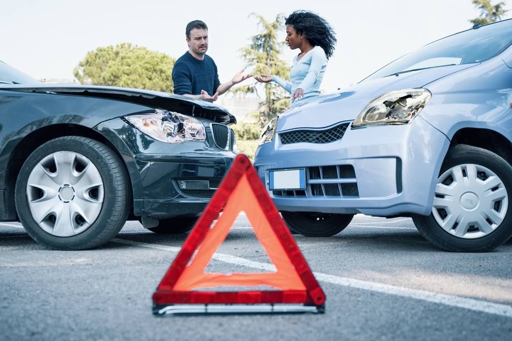 Two drivers arguing beside their damaged cars after a front-end collision, with a red warning triangle on the road in the foreground