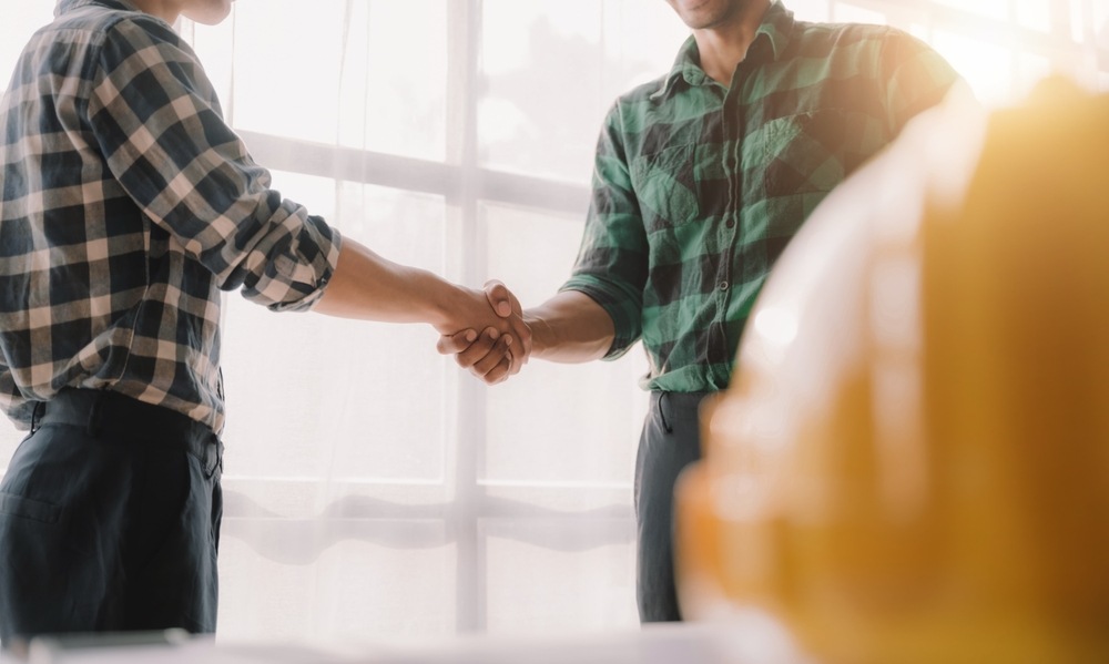 Two workers in plaid shirts shaking hands in a bright office, with a yellow hard hat blurred in the foreground