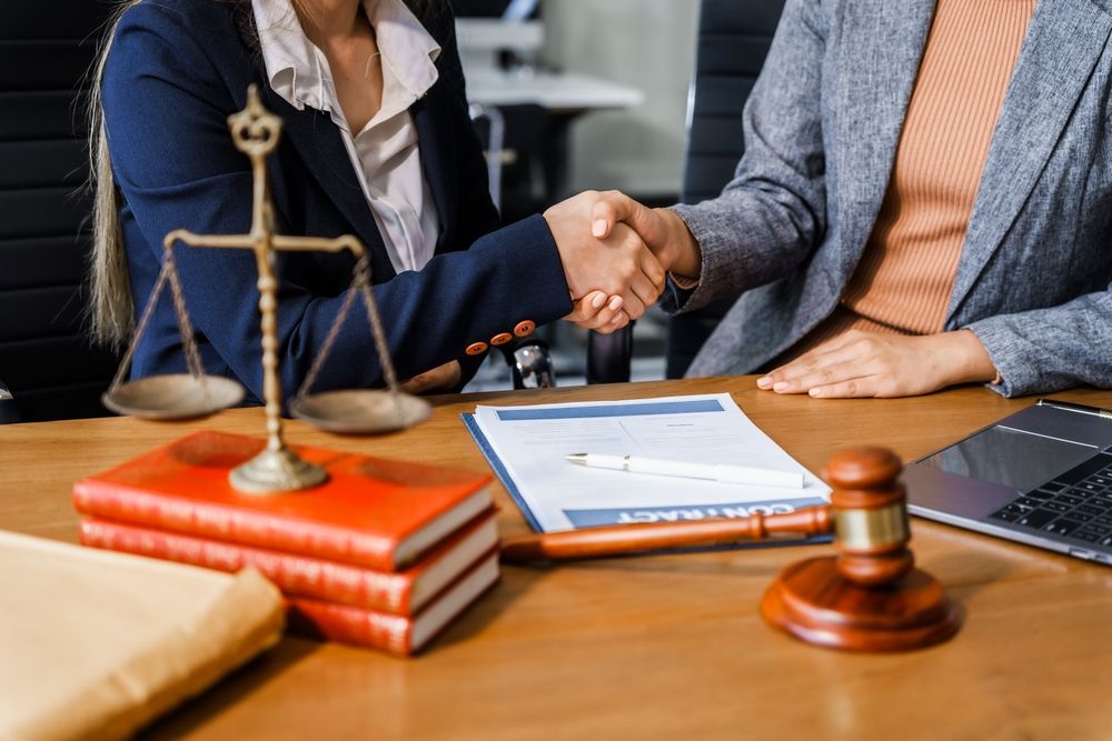 Two people shaking hands across a desk with a gavel, scales of justice, law books and a signed document