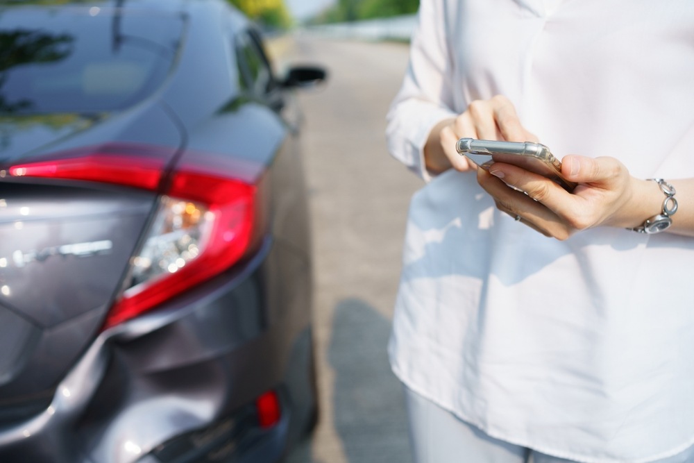 Driver holding a smartphone next to her car’s damaged rear bumper and taillight after a collision