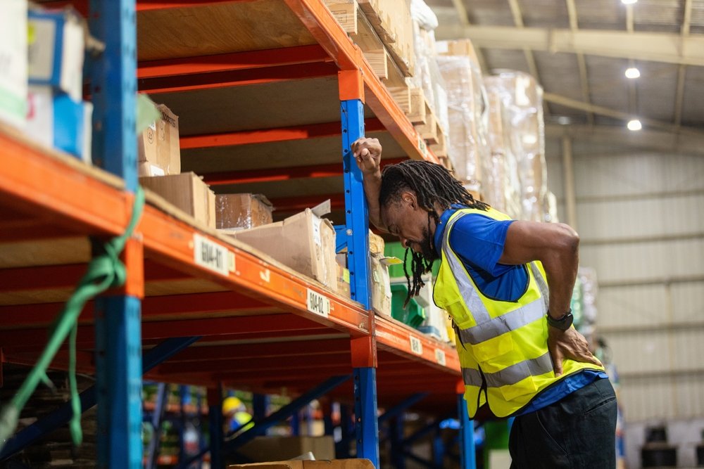 Warehouse worker in a high-visibility vest leaning on a storage rack and holding his lower back in pain