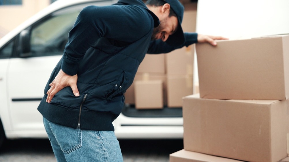 Delivery driver in a cap and jacket holding his lower back in pain beside a stack of boxes and an open delivery van