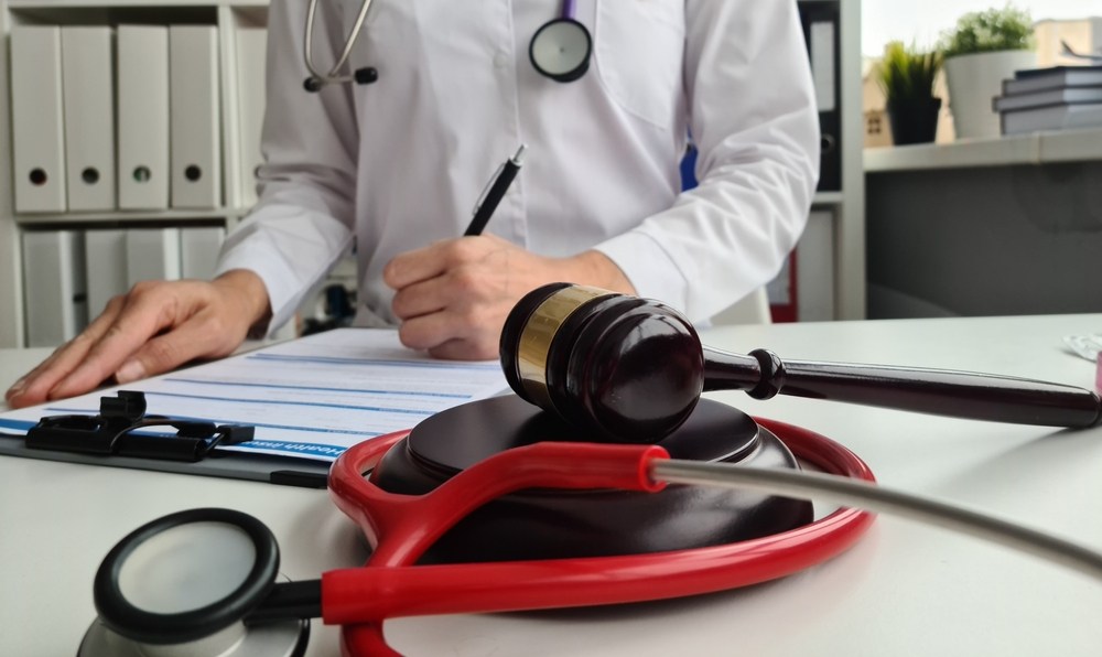 Doctor in a white coat filling out a medical form at a desk beside a gavel resting on a red stethoscope