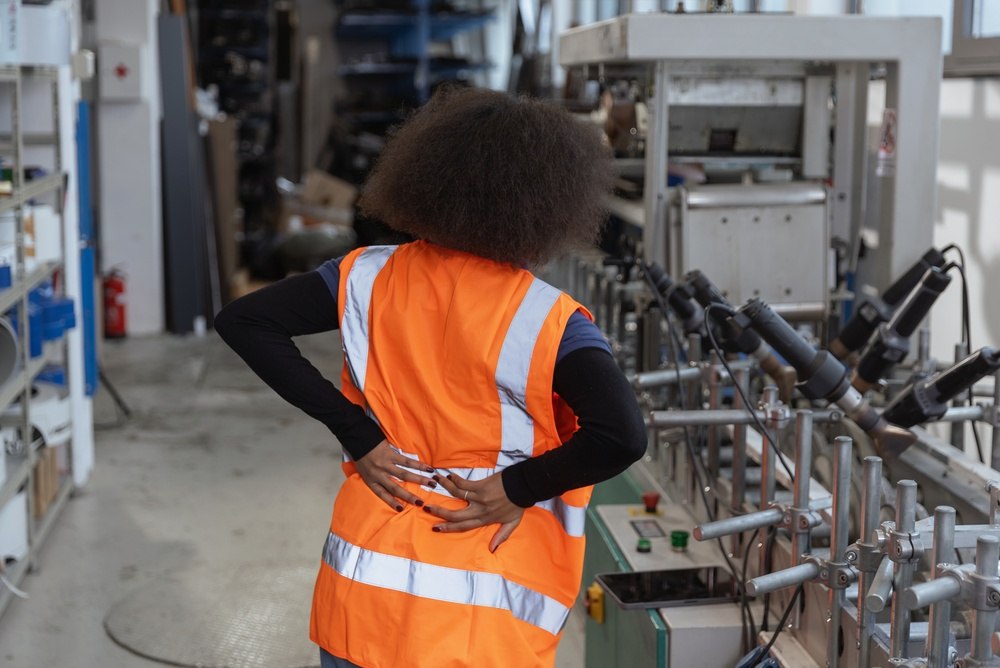 Factory worker in an orange safety vest holding her lower back while standing beside an industrial production line