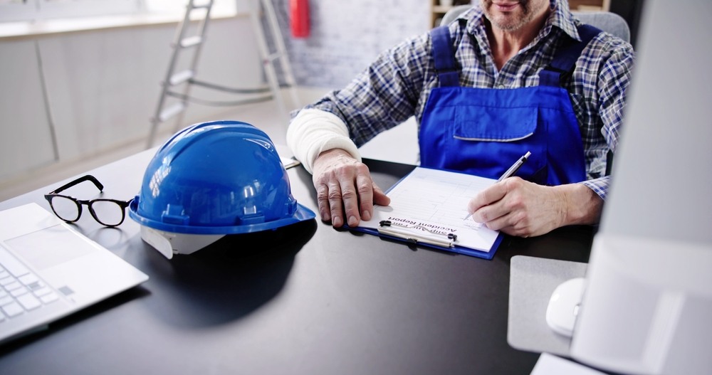 Worker in overalls with a bandaged forearm filling out an accident report at a desk beside a blue hard hat and a laptop
