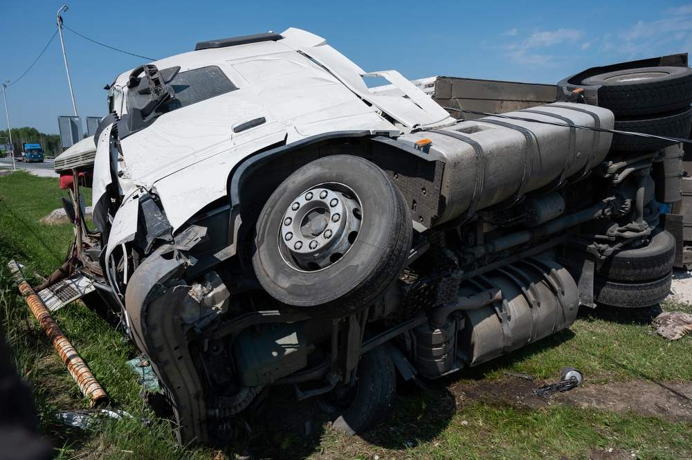 Overturned white semi truck cab lying on its side on a grassy roadside after a crash, with its fuel tank and undercarriage exposed