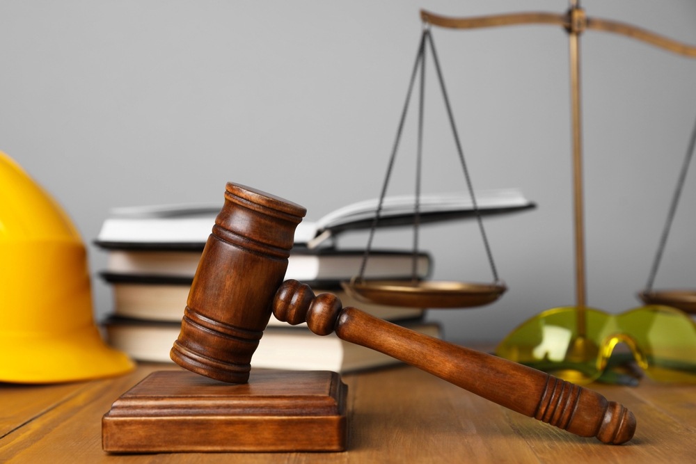 Wooden gavel and brass scales of justice on a desk beside a yellow hard hat, safety glasses, and a stack of law books