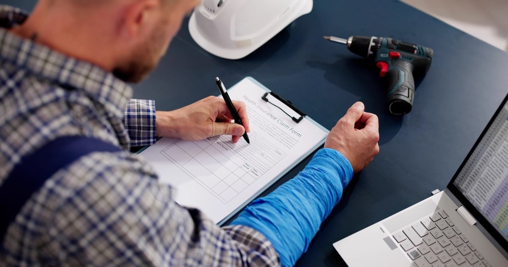 Injured worker with a wrapped forearm filling out a claim form at a desk beside a hard hat, a cordless drill, and a laptop