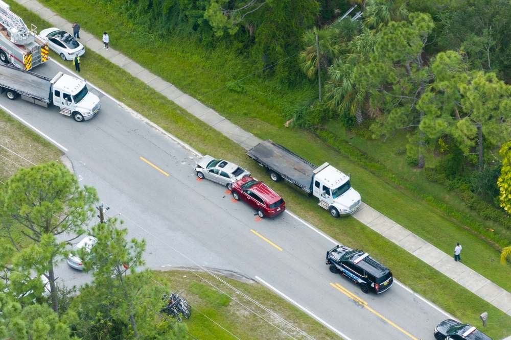 Aerial view of a rear-end collision on a two-lane road with evidence markers painted beside the cars, tow trucks and a police vehicle at the scene