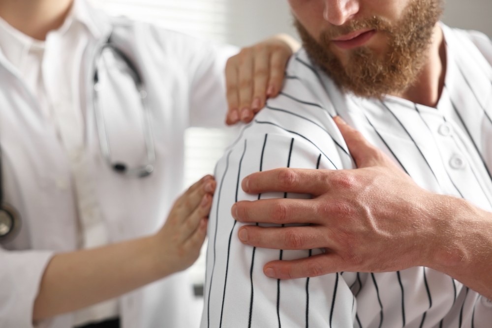 Physician examining a patient’s injured shoulder with both hands while the patient holds his upper arm